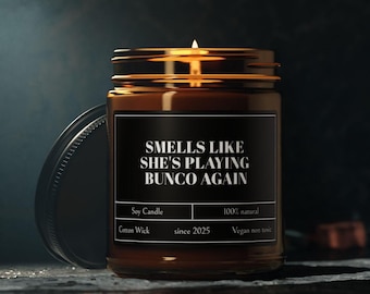 May include: A lit soy candle in a brown glass jar with a black label. The label reads "SMELLS LIKE SHE'S PLAYING BUNCO AGAIN." Additional text includes "Soy Candle," "Cotton Wick," "100% natural," "since 2025," and "Vegan non toxic."