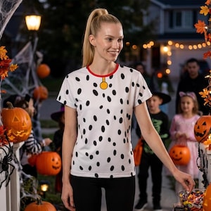 May include: A white short-sleeved t-shirt featuring a black dalmatian spot pattern, a red crew neckline, and a yellow circular tag graphic at the center of the collar, designed to resemble a dog costume.
