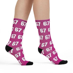 May include: Magenta socks patterned with the number "67" in white. The socks have black heels and toes. The socks are worn on a person's legs.