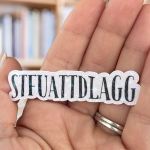 May include: A white sticker with a glittery background and black text that reads "STFUATTDLAGG".