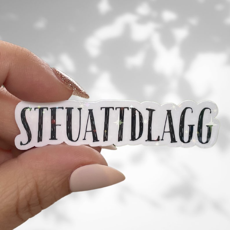 May include: A white sticker with black text that reads "STFUATTDLAGG" with a glittery background.