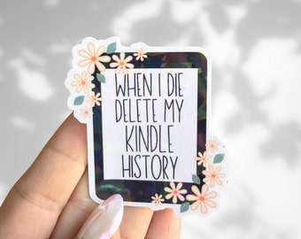 Puede incluir: Una pegatina con fondo negro y texto blanco que dice "Cuando muera, elimina mi historial de Kindle". La pegatina está rodeada de flores blancas con centros rosas.
