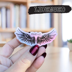 May include: A clear sticker with a pink and blue gradient background featuring a pair of black angel wings. The text "End of." is printed in black on the sticker.
