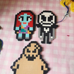 May include: Pixel art figures of Sally, Jack Skellington, and Oogie Boogie from The Nightmare Before Christmas. Sally has red hair, a blue face, and a patchwork dress. Jack is in a black suit. Oogie Boogie is tan.