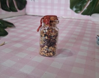May include: A small, clear glass bottle filled with dried herbs and flowers, sealed with a cork and a red wax drip. The bottle is set against a pink and white checkered background, suggesting a natural or apothecary theme.