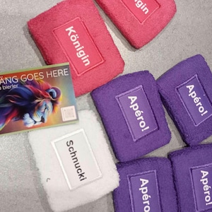 Könnte beinhalten: Eine Sammlung rechteckiger Frottee-Armbänder in Pink, Lila und Weiß, jeweils mit einem aufgenähten Etikett mit dem Text Königin, Apéro! oder Schnucki. Eine rechteckige Karte mit einer bunten Löwenillustration und dem Text DURÄÄNG GOES HERE by daniela bierler liegt bei.