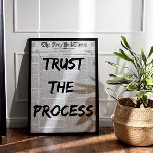 May include: A black and white framed print with the text "TRUST THE PROCESS" printed on a newspaper background. The print is hanging on a white wall.