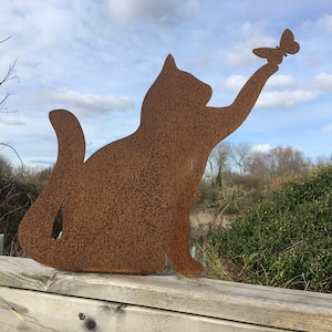 May include: A rusty metal silhouette of a cat reaching up to catch a butterfly. The cat is sitting on a wooden surface.