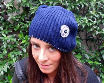 May include: A blue knitted beanie with a white stitched seam and a decorative eye-shaped button. The beanie is worn by a person with long brown hair.