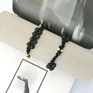 Puede incluir: Dos pulseras negras con cuentas marrones y blancas. Las pulseras están hechas de un cordón trenzado y tienen un diseño sencillo.