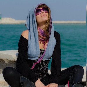 May include: A woman wearing a light blue hooded scarf with a pink and purple patterned scarf underneath. She is wearing sunglasses and looking up at the sky.