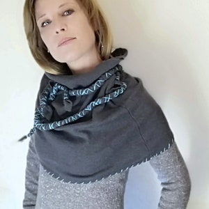 May include: A gray cowl neck sweater with a blue and gray braided trim. The sweater has a loose fit and a long, flowing design.