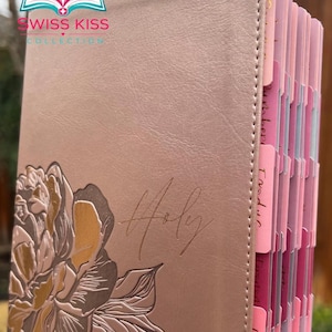 May include: A blush-colored Bible with a floral design and the word "Holy" embossed on the cover. The Bible has pink and white tabbed pages. The Swiss Kiss Collection logo is visible in the upper left corner.