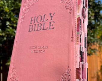 May include: A pink Holy Bible, King James Version, with decorative corner designs. The cover features the words "HOLY BIBLE" in silver lettering. The book is open, revealing colorful, floral-patterned page dividers.
