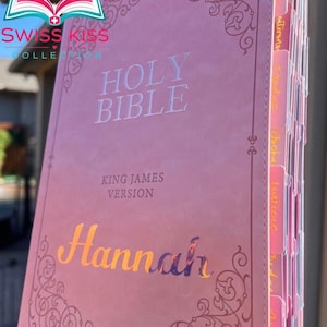 May include: A pink Holy Bible with the text "Holy Bible" and "King James Version" in white. The name "Hannah" is printed in gold foil. The cover has an ornate design. The book has pink and white tabs.