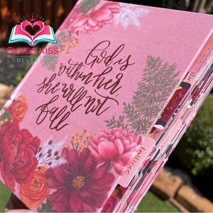 May include: A pink journal with floral designs and the text "God is within her she will not fall." The journal features a variety of flowers in shades of red, pink, and orange. The journal has labeled tabs.