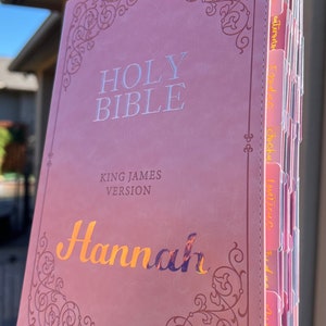 May include: A pink leather-bound Holy Bible with gold lettering. The Bible is personalized with the name "Hannah" in gold lettering. The Bible has tabs for each book of the Bible.