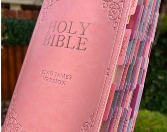 May include: A pink Holy Bible, King James Version, with decorative corner designs. The book has labeled tabs in various colors. The cover features the words "Holy Bible" in silver lettering. The Swiss Kiss Collection logo is visible in the bottom left corner.