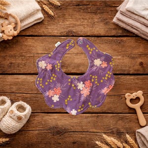 Puede incluir: Un babero morado para beb&eacute; con un estampado floral de flores rosas y blancas y detalles amarillos. El babero est&aacute; rodeado de art&iacute;culos para beb&eacute;s, incluyendo un par de zapatos de beb&eacute; de punto blanco, una estrella de madera y un mordedor de madera con forma de oso.