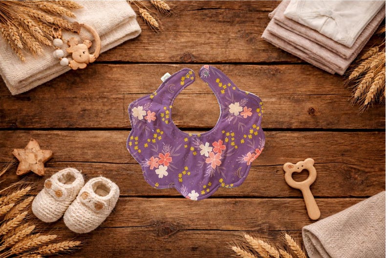 Puede incluir: Un babero morado para beb&eacute; con un estampado floral de flores rosas y blancas y detalles amarillos. El babero est&aacute; rodeado de art&iacute;culos para beb&eacute;s, incluyendo un par de zapatos de beb&eacute; de punto blanco, una estrella de madera y un mordedor de madera con forma de oso.