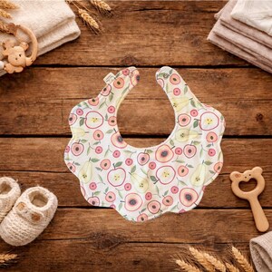 Puede incluir: Un babero para beb&eacute; con borde festoneado, estampado con manzanas y peras en tonos rosa, melocot&oacute;n y verde sobre fondo blanco. Rodeado de art&iacute;culos para beb&eacute;s, como patucos, una estrella de madera y un mordedor.