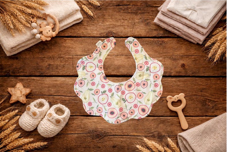 Puede incluir: Un babero para beb&eacute; con borde festoneado, estampado con manzanas y peras en tonos rosa, melocot&oacute;n y verde sobre fondo blanco. Rodeado de art&iacute;culos para beb&eacute;s, como patucos, una estrella de madera y un mordedor.