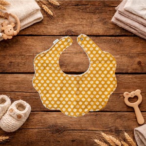 Puede incluir: Un babero amarillo con un estampado floral blanco es el punto focal de esta imagen. Est&aacute; rodeado de art&iacute;culos para beb&eacute;s, incluyendo botines de punto, una estrella de madera y un mordedor con forma de oso. Los art&iacute;culos est&aacute;n dispuestos sobre una superficie de madera r&uacute;stica.