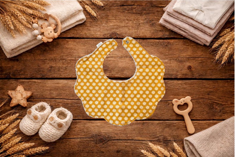 Puede incluir: Un babero amarillo con un estampado floral blanco es el punto focal de esta imagen. Est&aacute; rodeado de art&iacute;culos para beb&eacute;s, incluyendo botines de punto, una estrella de madera y un mordedor con forma de oso. Los art&iacute;culos est&aacute;n dispuestos sobre una superficie de madera r&uacute;stica.
