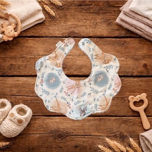 Puede incluir: Un babero para beb&eacute; con un dise&ntilde;o de animales del bosque, con ciervos y elementos florales en suaves colores pastel. El babero est&aacute; rodeado de art&iacute;culos para beb&eacute;s como patucos, una estrella de madera y un sonajero, todo sobre una superficie de madera.
