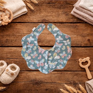 Puede incluir: Un babero para beb&eacute; con fondo azul y un estampado de mariposas de colores. El babero est&aacute; rodeado de art&iacute;culos para beb&eacute;s, incluyendo un par de botines de punto, una estrella de madera y un sonajero de madera, todo sobre una superficie de madera.