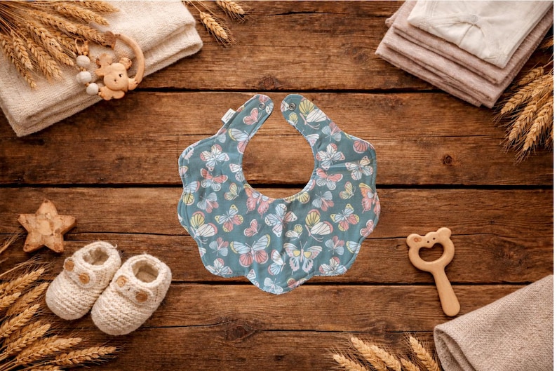 Puede incluir: Un babero para beb&eacute; con fondo azul y un estampado de mariposas de colores. El babero est&aacute; rodeado de art&iacute;culos para beb&eacute;s, incluyendo un par de botines de punto, una estrella de madera y un sonajero de madera, todo sobre una superficie de madera.