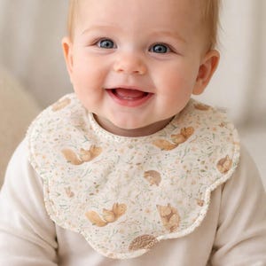 Puede incluir: Un beb&eacute; sonriente con un babero color crema con borde festoneado. El babero presenta un patr&oacute;n repetido de ardillas, erizos y elementos florales en tonos marrones y verdes. El beb&eacute; lleva una camisa de manga larga color crema.