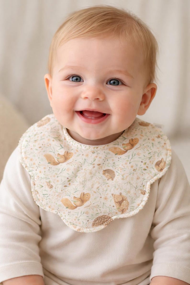 Puede incluir: Un beb&eacute; sonriente con un babero color crema con borde festoneado. El babero presenta un patr&oacute;n repetido de ardillas, erizos y elementos florales en tonos marrones y verdes. El beb&eacute; lleva una camisa de manga larga color crema.
