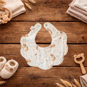 Puede incluir: Un babero para beb&eacute; con un estampado floral y de ardillas, zapatos de beb&eacute;, una estrella de madera y un mordedor con forma de oso. Los art&iacute;culos est&aacute;n dispuestos sobre una superficie de madera r&uacute;stica, con espigas de trigo y toallas dobladas.