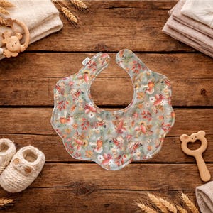 Puede incluir: Un babero para beb&eacute; con un estampado floral en tonos verdes, rosas y naranjas, rodeado de art&iacute;culos para beb&eacute;s sobre una superficie de madera. El babero es el punto focal, con un borde festoneado. Otros art&iacute;culos incluyen zapatos de beb&eacute;, una estrella de madera y un mordedor con forma de oso.