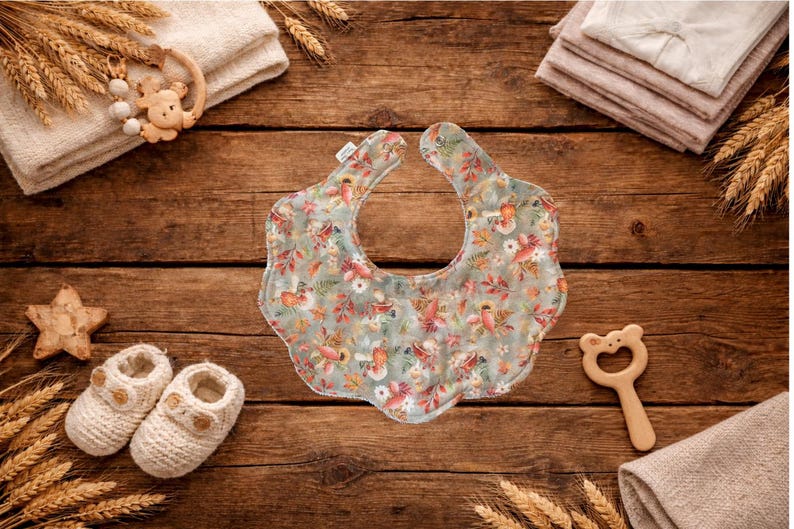 Puede incluir: Un babero para beb&eacute; con un estampado floral en tonos verdes, rosas y naranjas, rodeado de art&iacute;culos para beb&eacute;s sobre una superficie de madera. El babero es el punto focal, con un borde festoneado. Otros art&iacute;culos incluyen zapatos de beb&eacute;, una estrella de madera y un mordedor con forma de oso.