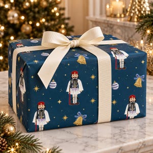 May include: A rectangular gift box wrapped in dark blue paper featuring a repeating pattern of nutcracker soldiers, gold bells with blue bows, striped ornaments, and yellow stars. The box is tied with a wide, cream-coloured fabric ribbon.