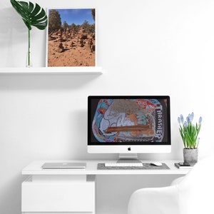 May include: A white desk with a white chair, a laptop, a desktop computer, a plant, and a framed photo of a desert landscape. The desktop computer has a screen displaying a colorful image with the text "Vans Off The Wall" and "Thrasher".