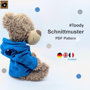 Peut inclure: Un ours en peluche marron portant un sweat à capuche bleu. L'image contient le texte "#Toody Schnittmuster PDF Pattern" et les drapeaux de l'Allemagne, du Royaume-Uni et de la France. L'arrière-plan est blanc.