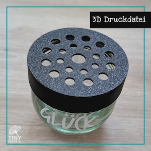 Può includere: Un barattolo di vetro trasparente con un coperchio nero perforato. Il coperchio ha un aspetto strutturato e glitterato. La parola "GLÜCK" è incisa in bianco sul barattolo. Il testo "3D Druckdatei" è in un rettangolo nero in alto a destra.