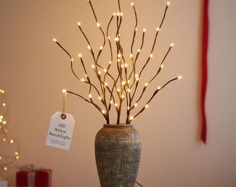 May include: LED willow branch lights in a textured gray vase. The branches are dark brown and adorned with small, warm white LED lights. A tag reads "LED Willow Branch Lights". The vase is on a white surface.