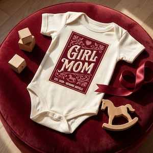 Puede incluir: Body de beb&eacute; color crema con un dise&ntilde;o rectangular burdeos que dice "Girl Mom" en escritura blanca. El dise&ntilde;o incluye un coraz&oacute;n, detalles florales y el texto "Est. 2026 - Genuine Article". Tambi&eacute;n hay juguetes de madera y una cinta.