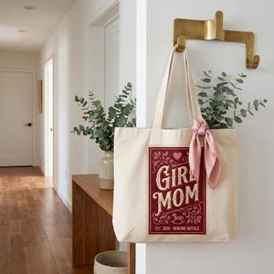 Puede incluir: Un bolso tote de color crema con un gr&aacute;fico burdeos que dice "Girl Mom" con elementos decorativos y el texto "EST. 2026 - GENUINE ARTICLE". La bolsa cuelga de un gancho de lat&oacute;n, con una bufanda rosa atada al asa.