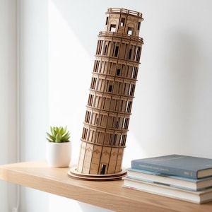Puede incluir: Maqueta de madera de la Torre de Pisa. El modelo es de madera marrón claro y está inclinado. Se exhibe en una estantería de madera con una pequeña planta en maceta y libros.