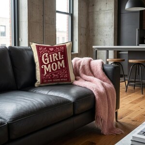 Puede incluir: Un coj&iacute;n decorativo crema con un borde burdeos y las palabras "GIRL MOM" en un dise&ntilde;o de estilo vintage. Una manta rosa est&aacute; sobre un sof&aacute; de cuero negro. El coj&iacute;n tambi&eacute;n tiene el texto "EST. 2028 - GENUINE ARTICLE".