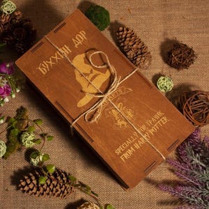 Op de afbeelding: Een houten kist met een scharnierend deksel, vastgebonden met touw. De kist heeft gegraveerde tekst en een silhouetontwerp. De tekst luidt "SPECIALLY FOR TRASHIK FROM HARRY POTTER".