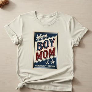 Puede incluir: Camiseta color crema con un gr&aacute;fico azul marino y rojo que dice "BOY MOM". El gr&aacute;fico incluye un tren, un ancla y una estrella. El texto "PREMIUM QUALITY - TRADEMARK" est&aacute; impreso debajo.
