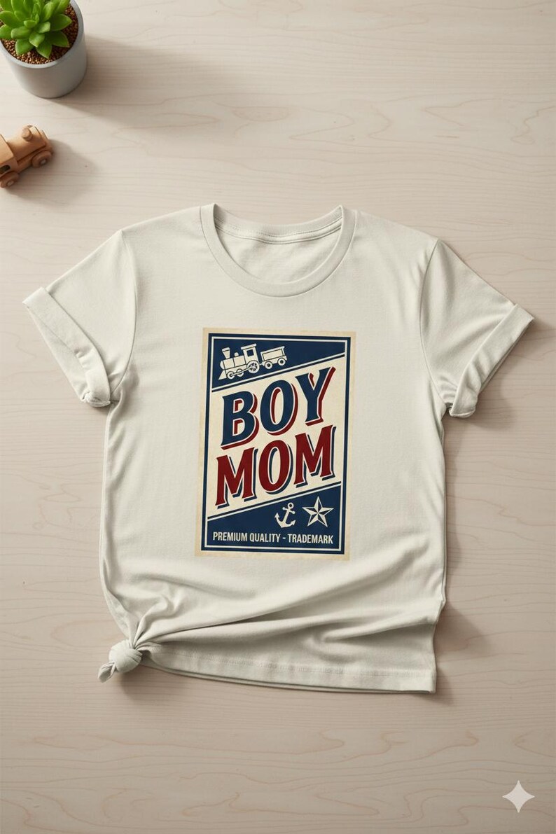 Puede incluir: Camiseta color crema con un gr&aacute;fico azul marino y rojo que dice "BOY MOM". El gr&aacute;fico incluye un tren, un ancla y una estrella. El texto "PREMIUM QUALITY - TRADEMARK" est&aacute; impreso debajo.