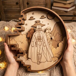 May include: A layered wooden artwork featuring Darth Vader and Stormtroopers from Star Wars. The circular design includes spaceships, clouds, and a planet. The artwork is held by hands and has the Etsy shop name "kreaco.etsy.com" at the bottom.