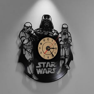 May include: Black Star Wars themed wall clock featuring Darth Vader and two Stormtroopers. The clock face is tan with black numbers and hands. The words "STAR WARS" are at the bottom of the clock.